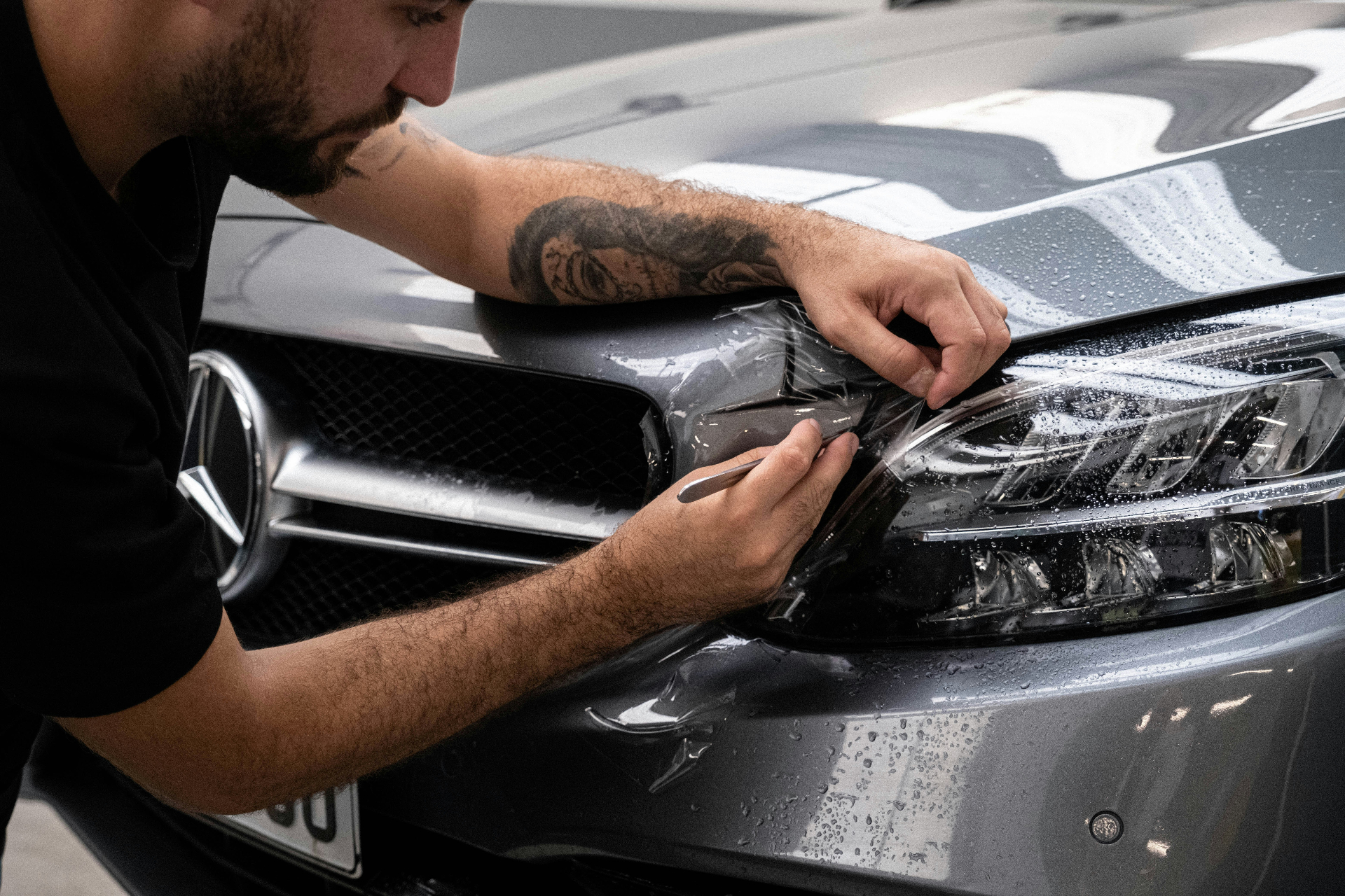 Detailer bringt vorsichtig eine transparente PPF-Lackschutzfolie am Scheinwerfer eines grauen Mercedes an